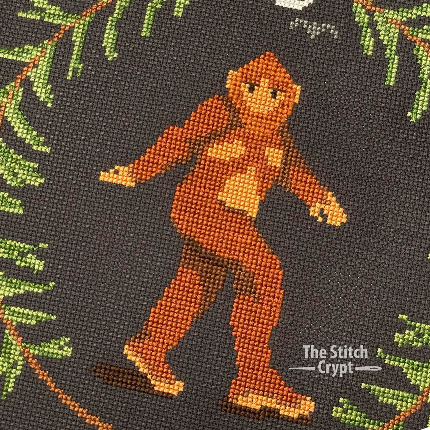 Striding Bigfoot centered inside a pine wreath with a skull at the bottom and a stylized half moon and bats at the top