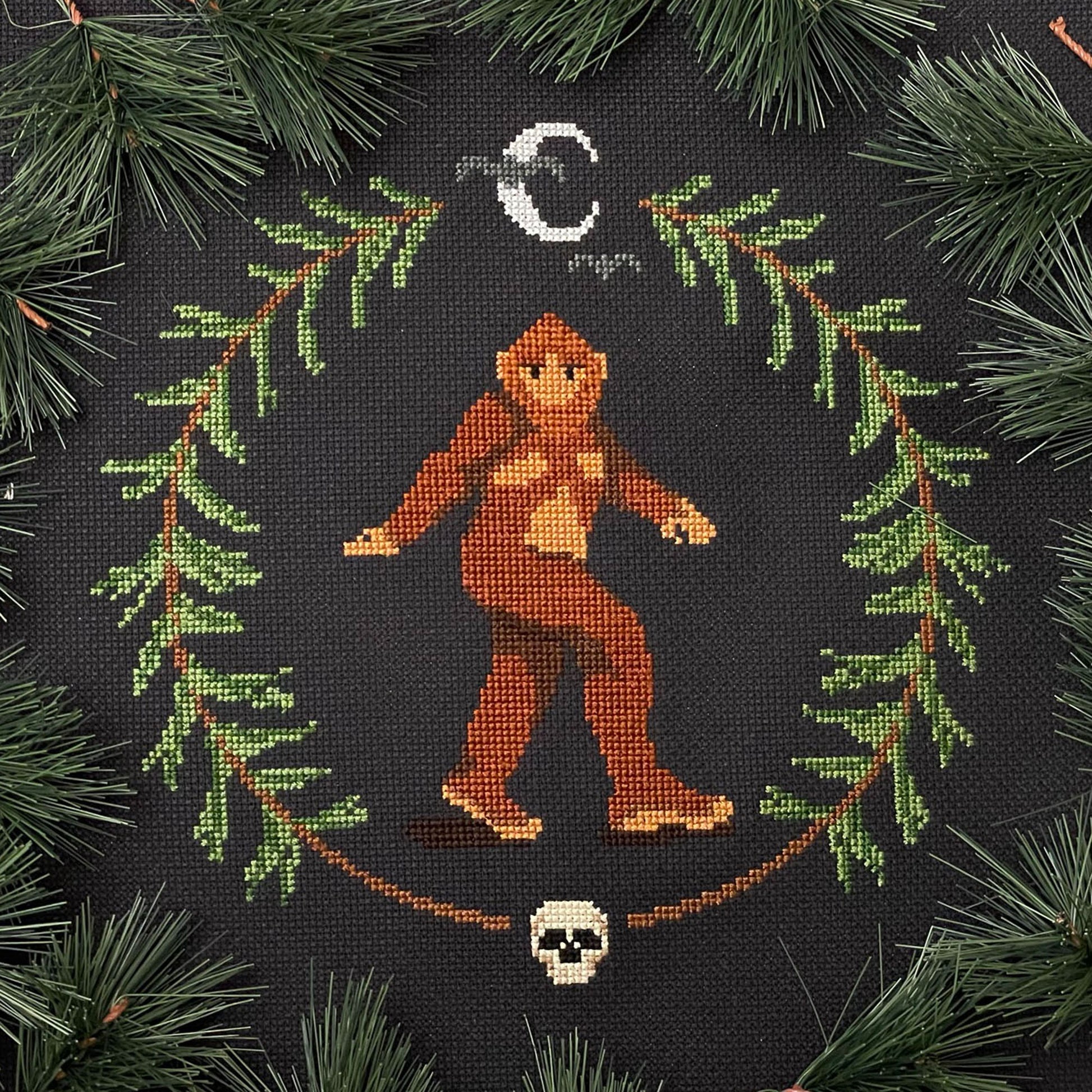 Striding Bigfoot centered inside a pine wreath with a skull at the bottom and a stylized half moon and bats at the top
